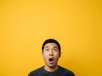 Surprised young man with dark hair and olive complexion wearing gray sweater against bright yellow background, expressing shock and amazement for advertising and marketing campaigns.