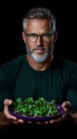 Mature man with gray hair and beard wearing glasses holds bowl of fresh microgreens against dark background. Professional food photography for healthy lifestyle concept.