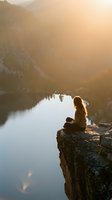 Young woman meditating on rocky cliff overlooking serene mountain lake at golden hour sunrise creating peaceful mindfulness moment in nature wilderness.