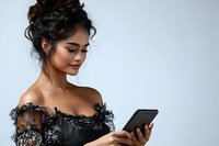 Young Asian woman in black lace dress looking at smartphone with gentle smile, messy updo hairstyle against light background. For lifestyle and technology content.