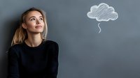 Young Caucasian woman in black sweater looking up thoughtfully at hand drawn cloud symbol on gray background, representing dreams, aspirations and creative thinking process.