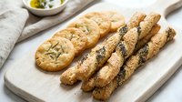 Freshly baked breadsticks with sesame and poppy seeds alongside crackers on wooden board, perfect for bakery, food styling, culinary blogs and restaurant menus.