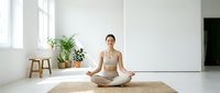Young woman of East Asian descent in athletic wear practicing yoga meditation in bright, minimalist studio space with plants and natural light for wellness and fitness content.