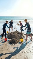 Four children in wetsuits building sandcastle on sunny beach with ocean waves. Kids playing together outdoors during summer vacation.