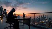 Professional woman working on laptop at sunset on high-rise balcony overlooking illuminated cityscape. Remote work, business, entrepreneurship.