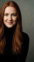 Young Caucasian woman with long red hair wearing black turtleneck sweater smiling warmly at camera against neutral gray background for professional headshot.