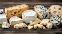 Assorted gourmet cheese varieties arranged on rustic wooden background, including blue cheese, brie, swiss and aged hard cheeses cut into pieces and chunks.