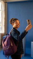 Happy young African American student woman taking selfie with smartphone wearing denim jacket and purple backpack in modern classroom setting.