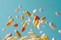 Various pharmaceutical pills, tablets, and capsules floating against turquoise background in dynamic composition, showing white, yellow and orange medication in motion.