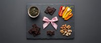 Elegant dark chocolate gift set with pink ribbon bow on slate board featuring nuts, candies and gourmet treats for special occasions and holidays.