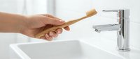 Hand holding eco-friendly bamboo toothbrush over modern bathroom sink with chrome faucet. Sustainable oral hygiene and zero waste lifestyle concept.