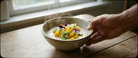 Person holding bowl of creamy soup with colorful vegetable garnish on wooden table by window, ready for lunch or dinner meal preparation and serving.