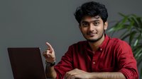 Young South Asian male professional in red casual shirt pointing upward while working on laptop, expressing positive attitude and confidence in modern office setting.