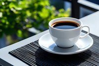 Fresh black coffee served in white porcelain cup and saucer on striped placemat, natural morning light through window with green foliage bokeh background.