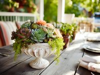 Colorful succulent arrangement in vintage stone urn on wooden table with blurred porch background, creating elegant outdoor dining atmosphere.
