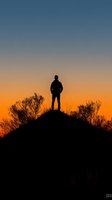 Silhouette of lone male figure standing on hilltop against dramatic orange and blue sunset sky, with bare branches framing the peaceful contemplative scene.