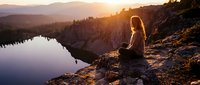 Young woman meditating at sunset by mountain lake, peaceful mindfulness practice in nature, golden hour light creating serene atmosphere for wellness and relaxation.