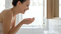 Young woman with olive skin tone rinsing face with water at bathroom sink, demonstrating skincare routine and morning hygiene habits for wellness and beauty content.