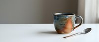 Handmade ceramic mug with colorful glaze beside a silver spoon on a white table, soft neutral background, cozy morning coffee concept.