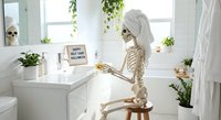 Skeleton with towel on head sits at modern bathroom vanity doing skincare routine surrounded by green plants in bright white space for Halloween humor.