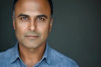 Professional headshot portrait of middle-aged South Asian man with intense gaze in blue shirt against dark background, perfect for business profiles.
