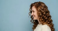 Young woman with curly red hair smiling in profile against blue background wearing cream sweater for lifestyle and beauty concepts.