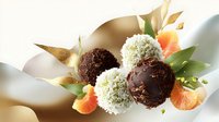 Dark chocolate truffles with white coconut coating arranged with orange citrus slices and green leaves on elegant golden background, luxury confectionery composition.