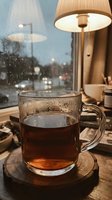 Glass mug of hot tea on wooden coaster by window with warm lamp light and rainy evening city view creating cozy cafe atmosphere.