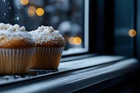 Freshly baked cupcakes dusted with powdered sugar sit on a windowsill with warm bokeh lights and snow falling outside, perfect for bakery, holiday, and food photography projects.