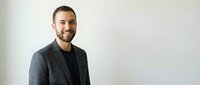 Young professional businessman with beard smiling confidently in modern office setting wearing casual blazer jacket against neutral background.