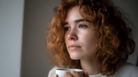 Young Caucasian woman with curly red hair and freckles gazing thoughtfully by window light, holding coffee cup in contemplative moment.
