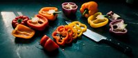 Fresh colorful bell peppers sliced and whole on dark surface with knife for healthy cooking and meal preparation in modern kitchen setting.