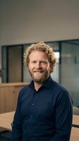 Confident middle-aged Caucasian businessman with curly blonde hair and beard smiling in modern office environment wearing navy blue shirt.