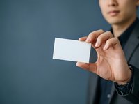 Young Asian male professional in dark business suit holding blank white business card against blue background, suitable for corporate branding and networking concepts.
