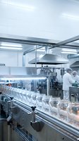 Modern pharmaceutical manufacturing facility with glass vials on automated production line and workers in sterile white uniforms operating equipment.