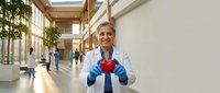 Female healthcare professional with blonde hair, Caucasian ethnicity, wearing white coat and blue gloves, holding red heart symbol in modern hospital corridor with natural light and blurred colleagues.