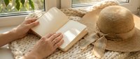 Woman reading open book by sunny window with straw hat and cozy knitted blanket creating peaceful morning atmosphere for relaxation and leisure time.