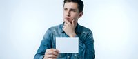 Young man with dark hair and light skin tone, wearing a blue denim shirt, holding blank white card while touching chin in thoughtful pose against white background.