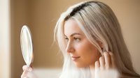 Young blonde woman examining her face in handheld mirror, touching skin with finger. Skincare routine, beauty treatment, self-care concept for cosmetics.