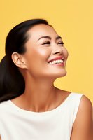 Young Asian woman with radiant smile and glowing skin wearing white sleeveless top against vibrant yellow background, expressing joy and confidence.
