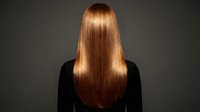 Woman with long straight shiny copper red hair from behind wearing black top against dark background for hair care and beauty concepts.