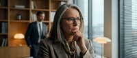 Middle-aged woman with long gray hair and glasses in professional business attire, contemplative expression with hand near face in modern office setting with colleague in background.