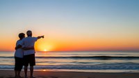 Couple embracing on beach at sunset watching golden hour over ocean waves with romantic sky colors reflecting on water for vacation memories.