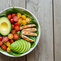 Healthy grilled chicken salad bowl with sliced avocado, cherry tomatoes and fresh greens on rustic wooden background for clean eating and nutrition.