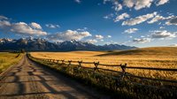Rural dirt road leading through golden prairie grassland toward snow-capped mountain range under dramatic blue sky with white clouds at sunset.