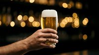 Male hand holding a glass of golden beer with foam head against blurred warm bokeh lights, suitable for beverage advertising and hospitality marketing.