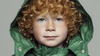 Young child with curly ginger hair wearing green winter jacket with hood, looking directly at camera against neutral background.