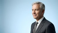 Professional mature Caucasian businessman with gray hair in dark suit smiling confidently against light blue background for corporate presentations.