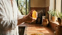 Man in white shirt squeezing fresh lemon juice over wooden counter with potted herbs, glass pitcher, and sunlit window in background for cooking and culinary content.