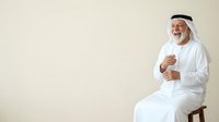 Middle-aged Middle Eastern man wearing traditional white thobe and ghutra, seated on wooden chair against neutral background, smiling warmly while gesturing with hands during conversation or presentation.
